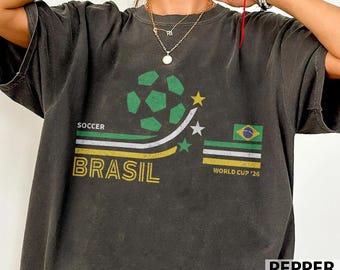 May include: Dark gray t-shirt with a soccer-themed graphic. The design includes a green and white soccer ball, the word "BRASIL" in yellow, and a Brazilian flag. The text "WORLD CUP '26" is also present.