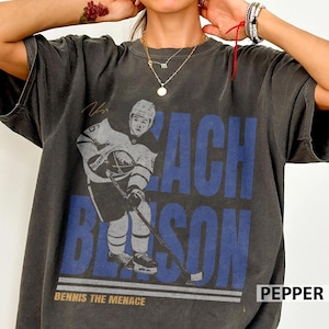 May include: Dark gray t-shirt featuring a hockey player graphic with the name "ZACH BENSON" in blue block letters. The text "BENNIS THE MENACE" is below the graphic. The shirt has a vintage, worn look.