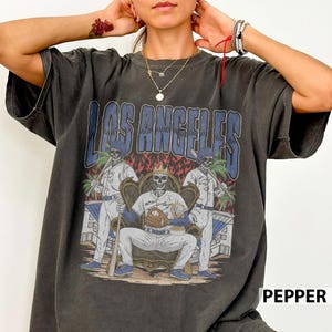 May include: Dark gray t-shirt featuring a graphic of skeleton baseball players with the text "LOS ANGELES" in blue. The design includes a skeleton seated on a throne holding a baseball and bat, with two other skeletons in the background.