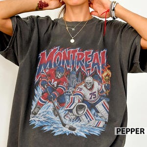 May include: A charcoal gray short-sleeve t-shirt featuring a graphic print of two skeleton hockey players. The text MONTREAL is printed above the players, who wear jerseys numbered 14 and 75. One player holds a hockey stick and the other a torch.