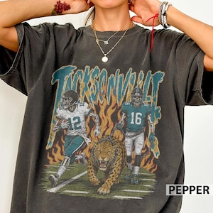 May include: A charcoal gray short-sleeve t-shirt featuring a graphic print of two football players with skeleton faces, one wearing number 12 and the other number 16, alongside a leopard, with orange flames and the text Jacksonville above.