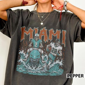 May include: A charcoal gray short-sleeve t-shirt featuring a graphic print of two skeleton football players. One player rides a dolphin in ocean waves, while the other runs with a football. The word MIAMI appears in orange above the figures.