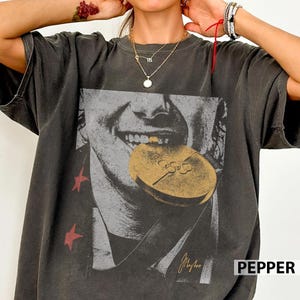 May include: Dark gray t-shirt featuring a black and white graphic of a person biting a gold medal. The medal has the Olympic rings symbol. Red star shapes are on the left side of the shirt. The word "PEPPER" is in white text on the bottom right.