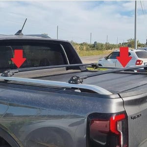 Puede incluir: Una camioneta gris oscuro con una cubierta de cama negra y una baca plateada. La baca tiene dos barras paralelas y está montada en la caja de la camioneta. La camioneta está estacionada al aire libre en un día soleado.