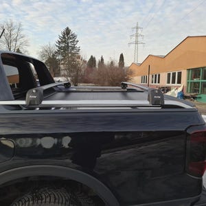 Puede incluir: Una caja de camioneta negra cuenta con una cubierta retráctil y barras transversales plateadas montadas en los rieles laterales. Los soportes de las barras llevan la inscripción PAV. El vehículo tiene un acabado negro brillante y luces traseras.