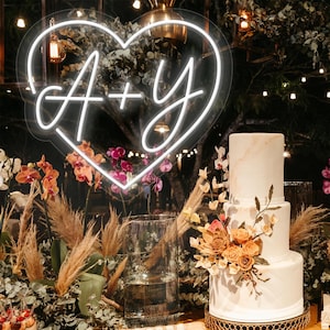 May include: A three-tiered white wedding cake with floral decorations sits on a gold stand. A neon sign in the shape of a heart with the letters "A + Y" is above the cake. The scene is decorated with flowers and greenery.
