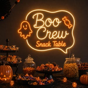 May include: A glowing orange neon sign shaped like a speech bubble with the text Boo Crew Snack Table, featuring a ghost and a candy graphic. The sign is mounted on a dark wall above a table filled with bowls of candy, popcorn, and pumpkins.