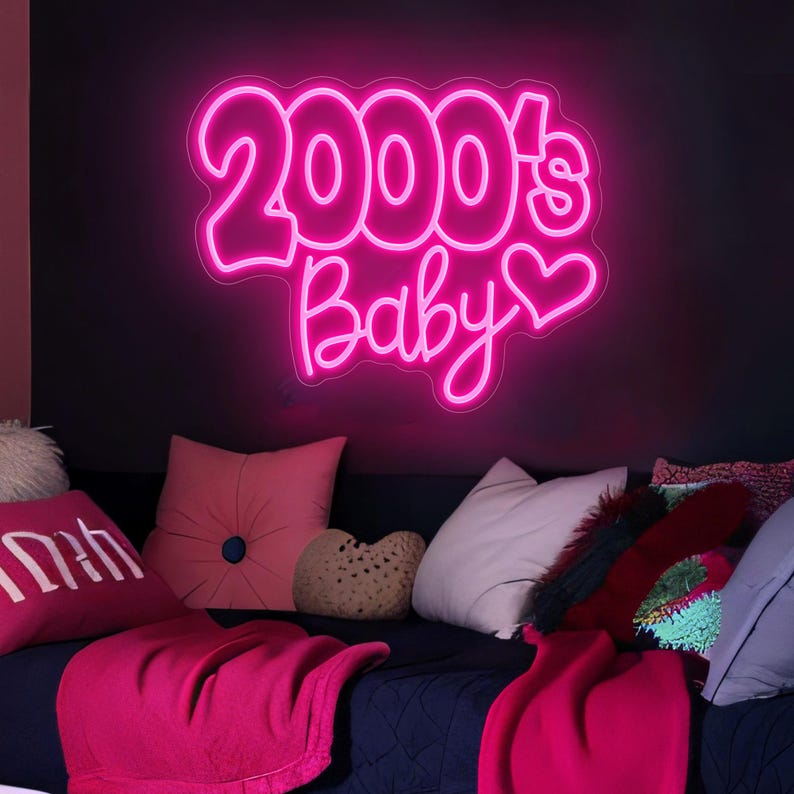 Pode incluir: Uma placa de n&eacute;on rosa brilhante exibe as palavras "2000's Baby" com um cora&ccedil;&atilde;o. Est&aacute; montada numa parede escura acima de uma cama adornada com almofadas rosa e brancas e uma manta rosa a condizer.