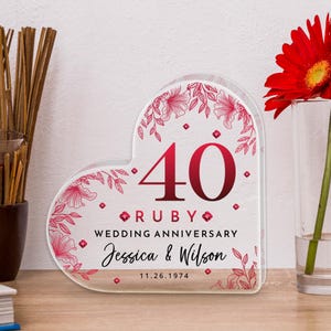 Può includere: Targa di anniversario a forma di cuore in acrilico trasparente con accenti floreali rossi. La targa recita "40° Anniversario di Matrimonio Rubino" con i nomi "Jessica & Wilson" e la data "11.26.1974". Una gerbera rossa in un vaso di vetro.