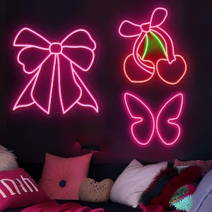 May include: Pink neon light fixtures in the shapes of a bow, cherries, and a butterfly are mounted on a dark wall. The cherries have a green stem. The room has pink and white pillows on a dark blue couch.