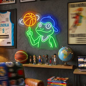 May include: A neon sign featuring a green frog wearing a blue baseball cap, spinning an orange basketball on its finger. The frog is outlined in bright green neon light, while the basketball is outlined in orange, creating a vibrant wall display.
