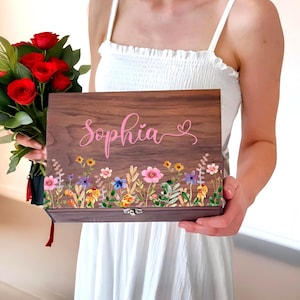 Könnte beinhalten: Eine braune Holzkiste mit floralem Design und dem Namen "Sophia" in rosa Schrift. Die Kiste hat einen Klappdeckel und einen Verschluss. Das florale Design umfasst rosa, gelbe, blaue und violette Blumen.