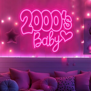 Pode incluir: Uma placa de n&eacute;on rosa brilhante com a frase "2000's Baby" e um cora&ccedil;&atilde;o. A placa est&aacute; montada numa parede rosa, com uma almofada em forma de estrela e luzes de corda. A cena &eacute; um quarto com almofadas rosas.