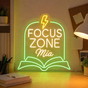 May include: A neon sign featuring a light green outline of an open book with a yellow lightning bolt icon above the text FOCUS ZONE Mia in white lettering. The sign is mounted on a clear acrylic backing and emits a bright glow on the desk surface.