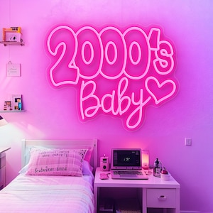 Pode incluir: Uma placa de n&eacute;on rosa que diz "2000's Baby" com um cora&ccedil;&atilde;o, montada numa parede rosa. O quarto apresenta uma cama com uma almofada rosa, uma secret&aacute;ria com um laptop e outros itens decorativos, tudo banhado em luz rosa.