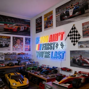 May include: A room decorated with racing-themed posters and model cars. A neon sign on the wall reads "IF YOU AIN'T FIRST YOU'RE LAST." A checkered flag hangs nearby. Various model race cars are displayed on shelves and tables.