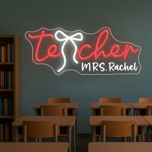 May include: A neon sign in a classroom setting. The sign reads "teacher" in red script with a white bow accent, and "MRS. Rachel" in white block letters. The sign is mounted on a teal wall above several student desks.