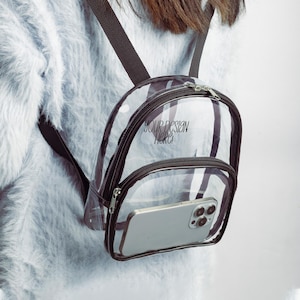 May include: A clear plastic mini backpack with dark brown trim, adjustable straps, and two zippered compartments. The front pocket contains a silver smartphone, and the text "YOUR DESIGN HERE!" is printed in black on the main compartment.