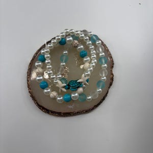 May include: Three beaded bracelets with a sea theme. The bracelets feature white faux pearls, turquoise beads, and clear beads. One bracelet has small white starfish charms and a turquoise turtle charm. The bracelets are displayed on a brown and tan decorative coaster.