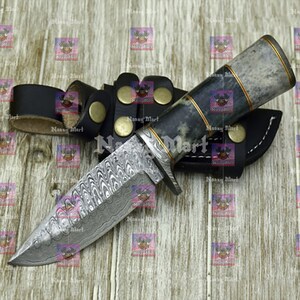 May include: A Damascus steel hunting knife with a patterned blade and a textured handle. The knife has a black leather sheath and a belt loop with brass-colored rivets. The handle is accented with gold-colored trim.