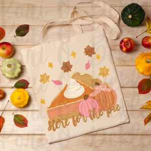 Peut inclure: Un sac fourre-tout beige avec un motif peint &agrave; la main d'une part de tarte &agrave; la citrouille avec de la cr&egrave;me fouett&eacute;e et deux citrouilles. Le texte "Here for the pie" est &eacute;crit en paillettes dor&eacute;es sur le sac.