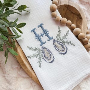 May include: White waffle-weave kitchen towel with blue embroidered letter "H" and oyster shell design. The towel is placed on a wooden tray with a strand of wooden beads and greenery.