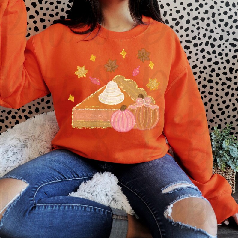 Peut inclure: Sweat-shirt orange avec un motif repr&eacute;sentant une part de tarte &agrave; la citrouille avec de la cr&egrave;me fouett&eacute;e et deux citrouilles. La tarte est d&eacute;cor&eacute;e de paillettes et de feuilles d'automne.