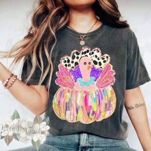 May include: A black t-shirt with a colorful graphic of a turkey inside a pumpkin. The turkey has pink feathers, a purple body, and a blue and white neck. The pumpkin is decorated with colorful stripes and glitter.