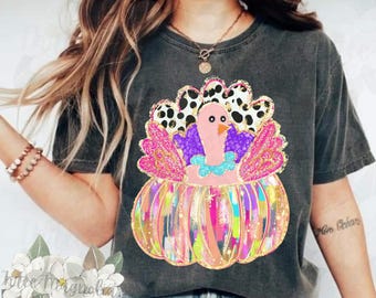 May include: A black t-shirt with a colorful graphic of a turkey inside a pumpkin. The turkey has pink feathers, a purple body, and a blue and white neck. The pumpkin is decorated with colorful stripes and glitter.
