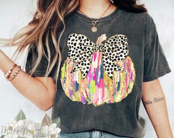 Peut inclure: Un t-shirt gris avec un motif de citrouille coloré. La citrouille a un nœud à pois noirs et blancs sur le dessus. Le t-shirt est fait d'un tissu doux et confortable.