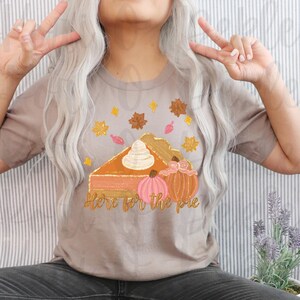 Peut inclure: Un t-shirt marron clair avec un motif repr&eacute;sentant une part de tarte &agrave; la citrouille avec de la cr&egrave;me fouett&eacute;e et deux citrouilles. Le texte "Here for the pie" est sous la tarte.