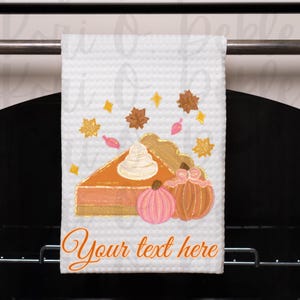 Peut inclure: Une serviette de cuisine blanche avec un motif peint &agrave; la main d'une part de tarte &agrave; la citrouille avec de la cr&egrave;me fouett&eacute;e et deux citrouilles. Le texte "Your text here" est imprim&eacute; sur la serviette.