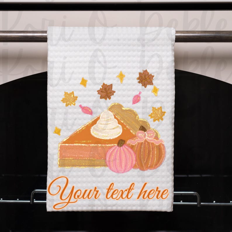 Peut inclure: Une serviette de cuisine blanche avec un motif peint &agrave; la main d'une part de tarte &agrave; la citrouille avec de la cr&egrave;me fouett&eacute;e et deux citrouilles. Le texte "Your text here" est imprim&eacute; sur la serviette.