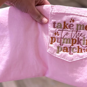 May include: A light pink sweatshirt with a pocket featuring the embroidered phrase "take me to the pumpkin patch" in shades of brown, orange, and pink. The pocket has a decorative stitched border.
