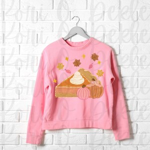 Peut inclure: Sweat-shirt rose avec un motif repr&eacute;sentant une part de tarte &agrave; la citrouille avec de la cr&egrave;me fouett&eacute;e et deux citrouilles. La tarte est d&eacute;cor&eacute;e de paillettes dor&eacute;es et de feuilles d'automne.