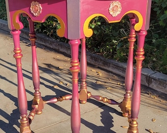 May include: A hand-painted, hexagonal side table in shades of pink and gold. The tabletop is a vibrant pink with a gold trim. The legs and decorative accents are painted in gold and pink, with floral embellishments.