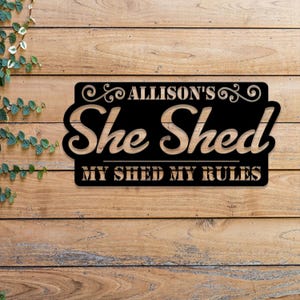 May include: Black metal sign with the text "Allison's She Shed - My Shed My Rules" on a wooden background.