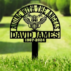May include: Black metal memorial garden stake with the text "Riding with the Angels" in a half-circle arch above the name "David James" and the dates "1967-2024". A motorcycle with wings is in the centre of the arch.