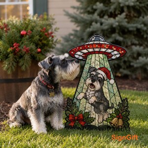 May include: A decorative yard sign featuring a stained-glass style illustration of a Schnauzer dog wearing a Santa hat, being beamed up by a UFO. The design includes festive holly, a red bow, and gold bells. The text SignGift is visible.