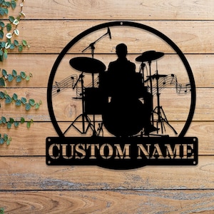 May include: Black metal silhouette of a drummer playing a drum set with the text "CUSTOM NAME" below. The silhouette is set against a woodgrain background.