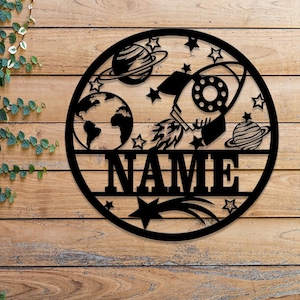 May include: Black metal wall decor with a space theme. It features a rocket, planets, stars, and a comet. The word "NAME" is in the center of the design.