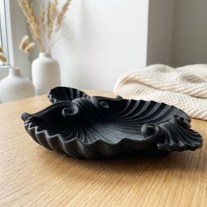 May include: A black, seashell-shaped decorative dish with a textured surface. The dish has a detailed, swirling design and sits on a wooden surface. Decorative vases and a cream-colored blanket are in the background.