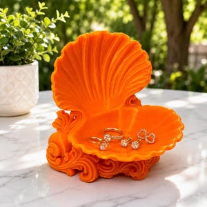 May include: A bright orange, clam-shaped jewelry dish featuring a scalloped shell design and an ornate, swirling base. The open shell holds several gold-toned rings and stud earrings with clear stones, resting on a smooth, white marble surface.