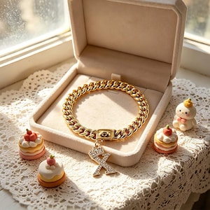 May include: A gold chain bracelet with an 'X' charm and crown, presented in a jewellery box. The bracelet is surrounded by miniature cakes and a small figurine on a lace surface. The bracelet's circumference is approximately 18 cm.