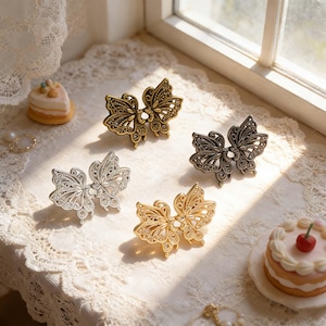 Pode incluir: Quatro objetos decorativos em forma de borboleta em tons de ouro, prata e bronze. Os desenhos de borboletas apresentam padrões intrincados e são adornados com pequenos detalhes brilhantes. Os objetos são exibidos em uma superfície coberta de renda.