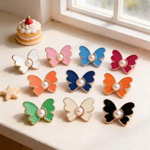 Puede incluir: Colección de broches de esmalte con forma de mariposa en varios colores, como blanco, rosa, azul, naranja, verde y negro. Cada broche tiene un contorno dorado y un centro de perla de imitación. También se ve un pequeño pastel y una decoración en forma de estrella.