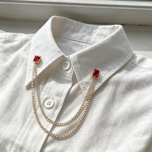 May include: A white button-down shirt featuring a gold-toned collar pin accessory. The accessory consists of two rectangular red stones set in gold frames, connected by two delicate gold-toned metal chains that drape across the shirt front.