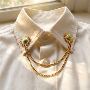 Puede incluir: Una camisa blanca de cuello rígido con un conjunto de broches dorados. El accesorio consta de dos piezas con forma de sol, cada una con una piedra turquesa lisa en el centro, unidas por dos delicadas cadenas doradas que cuelgan al frente.