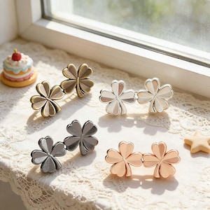 May include: Four pairs of clover-shaped hair clips in various metallic finishes: bronze, silver, gunmetal, and rose gold. Each clip features two four-leaf clovers linked together. A small cake and star-shaped decoration are also visible.