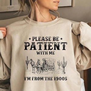 May include: A beige sweatshirt with a black graphic print that says "Please be patient with me I'm from the 1900s". The graphic also includes a black and white illustration of a horse-drawn wagon with cacti.